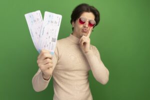 pleased young handsome guy wearing glasses holding tickets putting finger cheek isolated green wall min